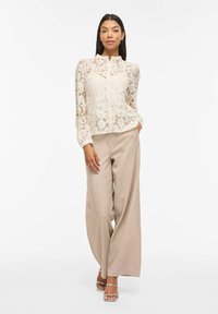 Blouse en dentelle blanc cassé avec manches longues et col montant, associée à un pantalon large beige clair et des sandales à talons transparents.