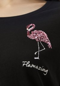 Rosa paljettflamingo med vita broderade ben och texten "Flamazing" på svart tyg, i närbild.