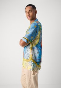 Camisa de manga corta y colorida con un patrón abstracto en azul, verde y amarillo, que presenta botones y un corte relajado. Llevada con pantalones beis.