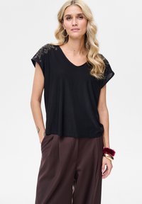 Czarny t-shirt z dekoltem w serek i krótkimi rękawami. Posiada ozdobne detale na ramionach. Noszony z brązowymi szerokimi spodniami. Gładka faktura materiału.