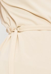 Μπεζ φόρεμα από ύφασμα με στενή ζώνη που κλείνει με δύο χρυσούς μεταλλικούς κρίκους, μαζεμένο στη μέση.