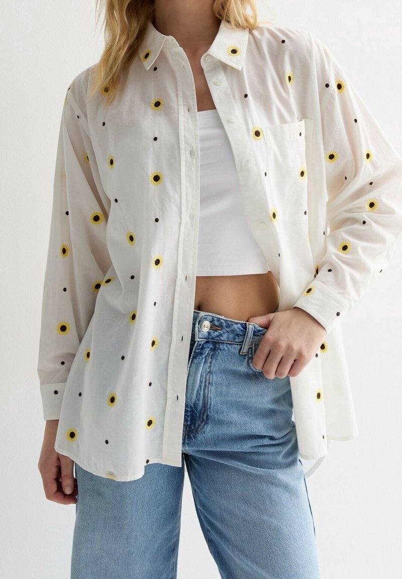 Femme portant une chemise blanche boutonnée avec de petits motifs jaunes de tournesols, un crop top blanc et un jean bleu taille haute.