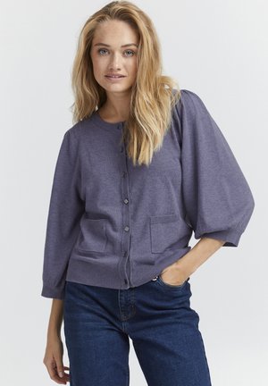 Paarse, lichte gebreide cardigan met een ronde halslijn, knopen aan de voorkant, twee zakken en ballonmouwen, gecombineerd met blauwe jeans.
