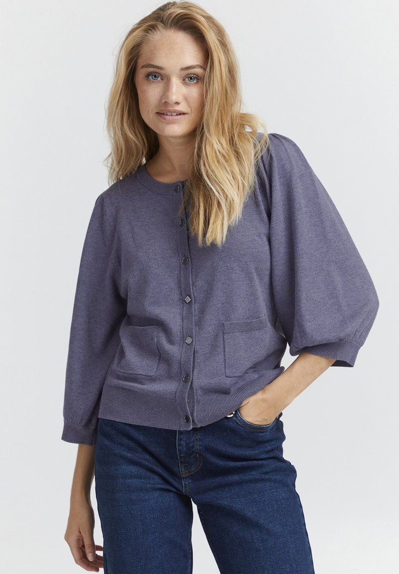 Lilla, kerge kootud kardigan ümmarguse kaeluse, ees olevate nuppudega, kahe tasku ja õhkrõhude varrukatega, koos siniste teksadega.