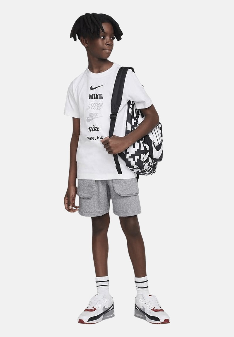 Camiseta gráfica de Nike en blanco con variaciones del logo en negro, pantalones cortos grises con bolsillos, mochila negra y blanca con estampado, y zapatillas Air Max.