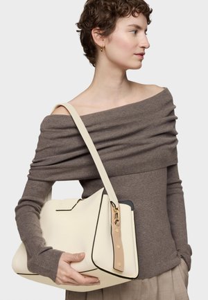 Mujer con un suéter marrón de hombros descubiertos que sostiene un gran bolso crema con una correa beige y herrajes en tono dorado.