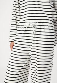 White pants with black horizontal stripes, featuring an elastic waistband with a drawstring. Soft fabric with a smooth texture.