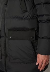 Giacca a vento nera con chiusura a zip, dotata di una grande tasca con bottoni a pressione, tessuto testurizzato e dettagli regolabili sul colletto.