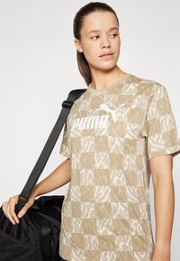 Beige kortermet t-skjorte med et subtilt dyremønster, med en hvit Puma-logo foran. Akkompagnert av en svart skulderveske.