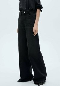 Jeans neri a gamba larga realizzati in denim, con vita alta, tasche large e orlo rifinito. Abbinati a una camicia scura con bottoni.