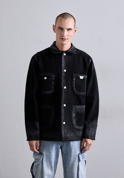 Homme portant une veste noire à boutons avec quatre poches avant et un jean cargo bleu clair, se tenant devant un mur gris clair uni.