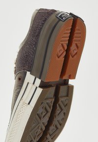 Zapatilla de lona marrón y gris con parte superior texturizada, suela de goma contrastante, parche con logo negro y detalles de franjas blancas en el lateral.
