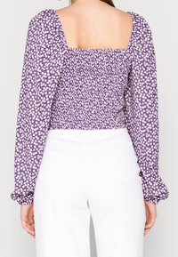 Fialová květinová blůza s dlouhými nafouklými rukávy, čtveratým výstřihem a nařasenou texturou. Obsahuje bílé květy na fialovém pozadí. Spárováno s bílými kalhotami.