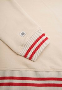 Beige sweatshirt med ribbede manchetter, der har røde og hvide striber. En mærkeetiket er fastgjort nær manchetten. Blød tekstur og afslappet design.