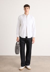 Witte overhemDB, zwarte broek, grijze handtas en witte schoenen met beige zool. Model poseert op een lichtgrijze vloer tegen een effen achtergrond.