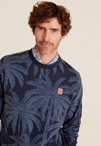 Sweatshirt marine avec un motif de feuilles de palmier en bleu clair, comportant une étiquette rectangulaire rouge sur la poitrine ainsi qu'un col et des poignets côtelés.