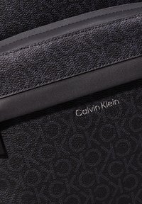 La borsa Calvin Klein presenta un materiale nero testurizzato con un motivo a logo ripetuto e una bordatura nera liscia. Inclusa un'accento con il nome del marchio in argento.
