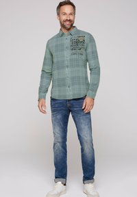 Camisa de cuadros verdes con un logo en el pecho, combinada con jeans azules y zapatillas blancas. La camisa tiene mangas largas y un dobladillo redondeado.