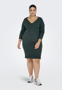 Robe tricotée verte avec un décolleté en V, des manches trois-quarts et une silhouette ajustée, assortie à des baskets de sport blanches et argentées.