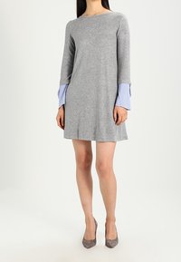 Grå stickad klänning med långa ärmar som har blå och vita randiga manschetter. A-linjeform, rund hals, mjuk textur, knälång.