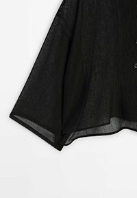 Manche de chemise en tissu noir léger semi-transparent avec une coupe ample et une patte de boutonnage visible sur fond blanc.