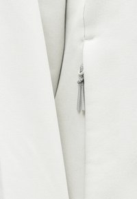 Tissu blanc texturé avec une fermeture éclair visible, comportant une fine tirette grise. Le design est épuré avec des lignes nettes et des accents minimes.