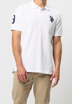 Mężczyzna ubrany w białą koszulkę polo z czarnym logo gracza polo na lewej piersi oraz granatową cyfrą 3 na prawej rękawie, w połączeniu z beżowymi spodniami.