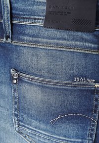 Blå denimjeans med en bakficka med en knapp, kontrasterande sömmar och en läderetikett präglad med "RAW 3301" och varumärket.