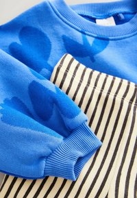 Blaues Sweatshirt mit abstraktem Muster und gerippten Bündchen, kombiniert mit gestreiften, beigen und dunkelbraunen Hosen. Der Stoff scheint weich mit unterschiedlichen Texturen zu sein.