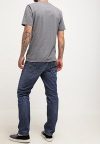 Uomo con una maglietta grigia, jeans blu e scarpe slip-on nere, con tatuaggi su entrambe le braccia, in piedi contro uno sfondo chiaro e semplice.