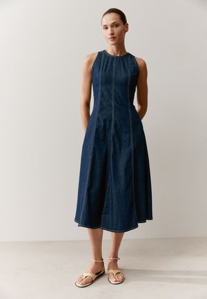 Frau steht vor einer schlichten Wand, trägt ein ärmelloses dunkles Denim-Midikleid und beige flache Sandalen mit Knöchelriemen, die Hände hinter dem Rücken.