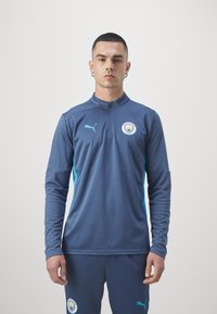 Temno modra atletska majica z zadrgo pri ovratniku, z svetloblue akcenti in logotipom Manchester City na prsih. Tekstura tkanine.