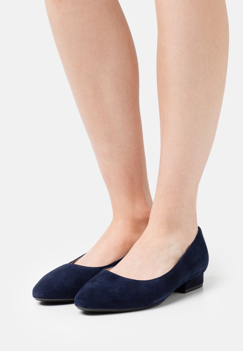 Jambes portant des chaussures fermées en daim bleu marine à talons bas sur un fond clair.