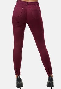 Jeans skinny burdeos con una textura suave, cintura alta, bolsillos traseros y detalles de dobladillo desflecado. Combinados con tacones altos negros.