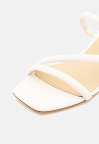 Sandale blanche à bout ouvert avec deux fines brides sur le devant et une bride à la cheville sur une semelle beige sur un fond uni.