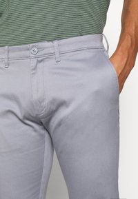 Světlé šedé bavlněné kalhoty s rovným předním dílem, bočními kapsami a zapínáním na knoflík. Mají hladkou texturu a rovný střih nohavic.