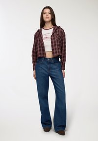 Camicia di flanella a quadri bordeaux tagliata, indossata sopra una t-shirt bianca con stampa, abbinata a jeans blu a gamba larga e scarpe marroni.