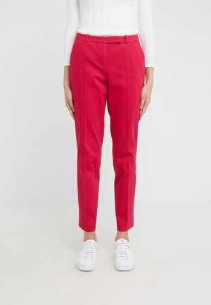 Femme portant un pantalon slim chevilles rouge vif et des baskets blanches, debout devant un fond clair uni.