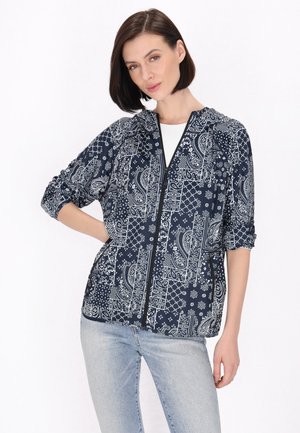 Marineblauwe jas met witte paisley- en geometrische patronen. Heeft een frontale rits, driekwart mouwen en een ontspannen pasvorm.