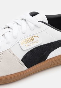 Bílá sneaker s černými a šedými semišovými akcenty, s zlatou logo nášivkou "Puma Palermo", bílými tkaničkami a texturovanou gumovou podrážkou.
