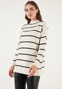 Maglione lungo beige chiaro a maniche lunghe con strisce orizzontali nere, caratterizzato da un collo alto e fessure laterali. Tessuto morbido, vestibilità comoda.