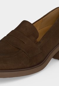 Mocassin en daim marron avec un petit talon, bout arrondi et détail de bride sur le dessus, montrant la doublure intérieure beige.