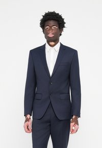Costum bleumarin cu două butoane, rever în formă de notch și pantaloni cu față dreaptă; asortat cu o cămașă albă cu guler. Țesătură netedă și design structurat.