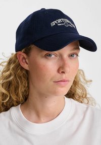 Ung kvinna med lockigt blont hår som bär en marinblå keps med texten "SPORTSWEAR" och en vit t-shirt med rund hals mot en vit bakgrund.