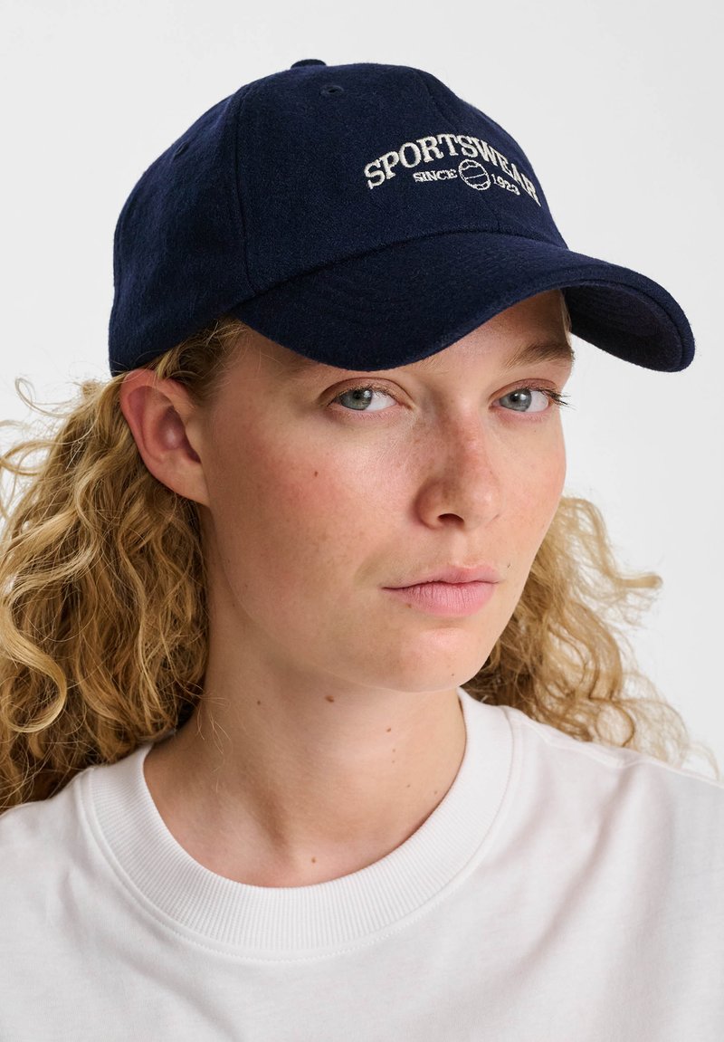 Ung kvinna med lockigt blont hår som bär en marinblå keps med texten "SPORTSWEAR" och en vit t-shirt med rund hals mot en vit bakgrund.