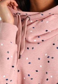 Felpa rosa realizzata in tessuto morbido, con collo a cowl, lacci e un motivo geometrico di quadrati blu navy, grigio e bianco.