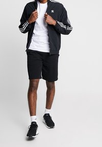 Giacca a vento nera con strisce bianche e logo, abbinata a pantaloni corti neri e scarpe da ginnastica nere. T-shirt bianca sotto. Outfit sportivo casual.