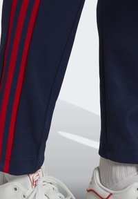 Marina blå träningsbyxor med röda ränder, tillverkade av ett slätt tyg. Har ribbade muddar och en nära passform, ihop med vita träningssneakers.
