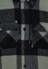 Veste en laine à grands carreaux noirs et gris, avec quatre boutons foncés, deux poches poitrine à rabat et cordons noirs.