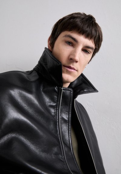 Veste en cuir noir avec un col haut, présentant une finition brillante et une fermeture éclair. Texture lisse sans motifs ni accents visibles.